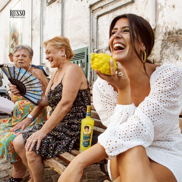 Limoncello d’Amalfi IGP Distillatori Russo in primo piano, incorniciato da vedute della costa e abitanti locali che celebrano la tradizione.