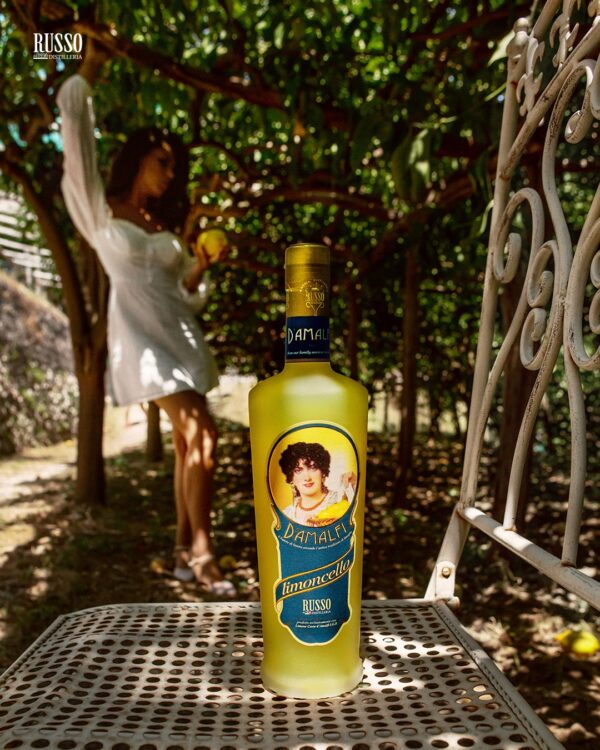 Limoncello d’Amalfi IGP Distillatori Russo in primo piano, incorniciato da vedute della costa e abitanti locali che celebrano la tradizione.